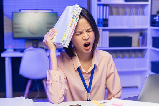 Stressed young Asian businesswoman overwhelmed with workload and holding documents while screaming in office.
