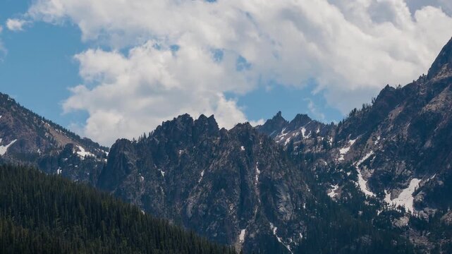 View of the jagged Sawtooth Mountains with snow patches and dense pine forests under a bright sky in Stanley, Idaho, United States.