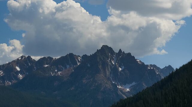 View of the jagged peaks of the Sawtooth Mountains with patches of snow under a bright blue sky with large white clouds in Stanley, Idaho, United States.