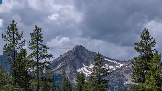 View of the rugged Sawtooth Mountains peak with patches of snow and evergreen pine trees under a dramatic cloudy sky in Stanley, Idaho, United States.