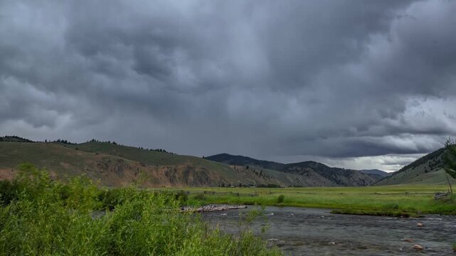 View of the Salmon River flowing through a lush green valley with rolling mountains under a dramatic stormy sky in Stanley, Idaho, United States.