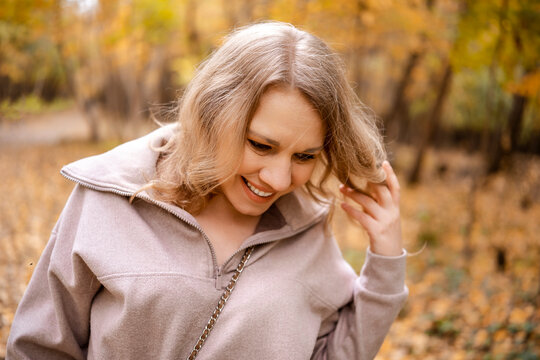 Laughing woman looking down in autumn park &mdash; spontaneous authentic happiness. Positive psychology and mental vitality boost. Carefree lady touching hair spending leisure time walking outside.