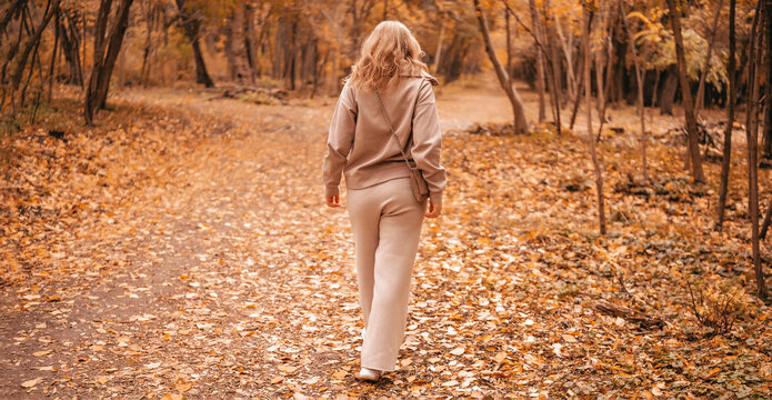 Woman walking away on autumn forest path &mdash; solo journey and self discovery. Life path philosophy and emotional independence. Adult lady strolling along yellow trees enjoying quiet outdoor leisure.