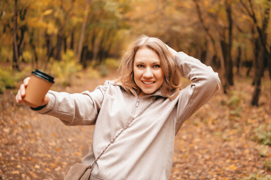 Joyful woman blogger showing coffee cup in autumn forest. Smiling tourist in beige sweater holding hot beverage among yellow leaves. Concept of warming up outdoors.