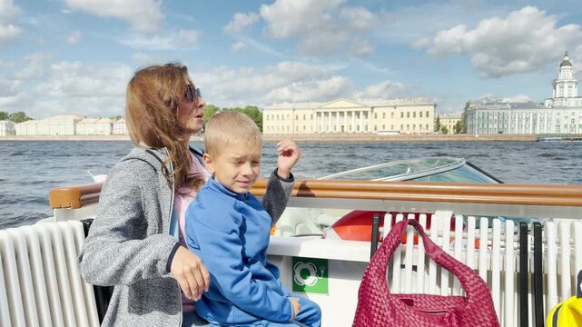 A young boy points eagerly from a boat deck in Saint Petersburg. His mother holds him with loving care across the wide river. Their joyful journey brings warmth and wonder.