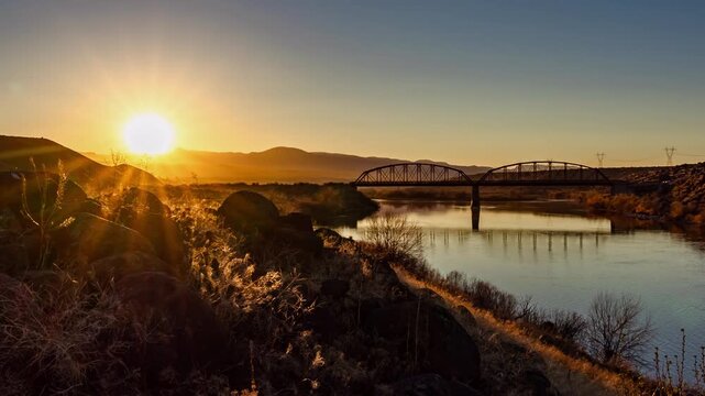 View of a steel truss bridge spanning a calm river at sunset with rocky terrain and distant mountains in Idaho, United States.