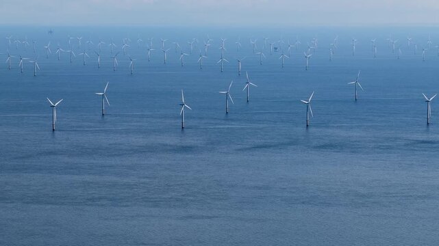 Aerial footage flying towards a vast offshore wind farm with rows of wind turbines standing across the open sea creating a striking renewable energy landscape stretching into the distance