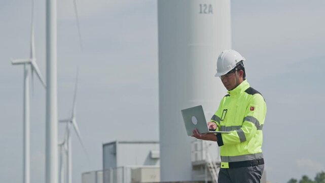 Engineer wearing safety helmet and reflective jacket using laptop during wind turbine maintenance, renewable energy technician inspecting equipment and monitoring outdoor facility operations