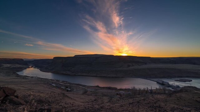 View of sunset over the Snake River canyon with a hydroelectric dam in the distance, featuring rugged cliffs and a vibrant sky in Idaho, United States.