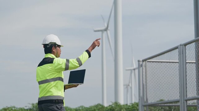 Engineer inspecting wind turbine equipment at renewable energy facility using laptop, wearing safety helmet and reflective vest during outdoor maintenance at fenced wind farm under cloudy sky