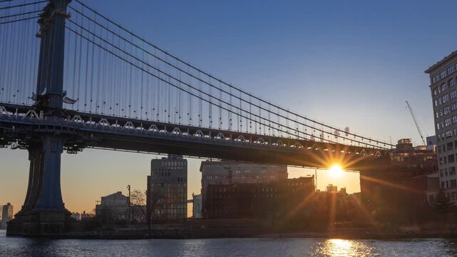 View of the Manhattan Bridge at sunset with the sun shining through the bridge structure, reflecting on the water, with city buildings in New York, New York, United States.
