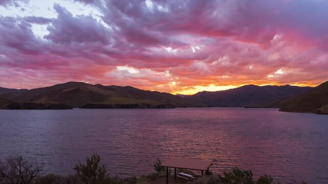 View of the calm waters of Lucky Peak Lake at sunset with a picnic shelter in the foreground and rolling hills under a vibrant purple and pink sky in Ada County, Idaho, United States.
