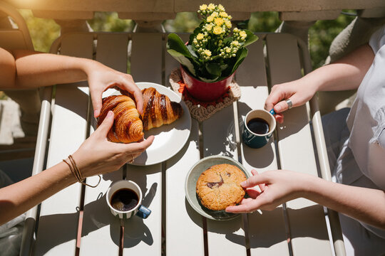 Friends sharing Croissants and Chocolate Cookie Beside Cups of Black Coffee and Yellow Flowers on a Sunny Outdoor Table. Summer coffee at sunny terrace.