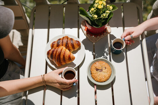 Coffee with friend at sunny terrace. Hands Holding Coffee Cups Beside Croissants, Chocolate Cookie and Yellow Flowers on a Sunny Outdoor Table