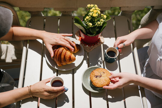 Friends sharing Croissants and Chocolate Cookie Beside Cups of Black Coffee and Yellow Flowers on a Sunny Outdoor Table. Summer coffee brake