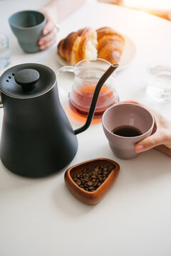 Hands Holding Cups of Fresh Black Coffee at a Morning Table with Gooseneck Kettle, Coffee Beans, Glass Server and Croissants. vertical composition