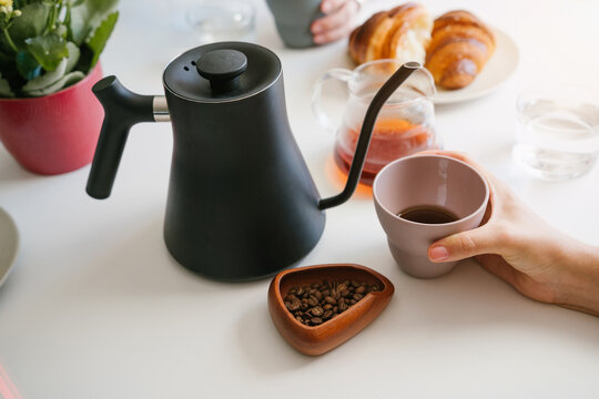 Hands Holding Cups of Fresh Black Coffee at a Morning Table with Gooseneck Kettle, Coffee Beans, Glass Server and Croissants. horizontal composition