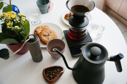 Manual V60 pour over coffee brewing with paper filter, fresh ground coffee, hand grinder, gooseneck kettle, roasted beans, glass server, cup, cookies and flowers on a white table at home