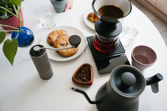 Manual pour over coffee brewing setup with V60 dripper, paper filter, freshly ground coffee, hand grinder, gooseneck kettle, roasted beans, glass server and cookies on white table