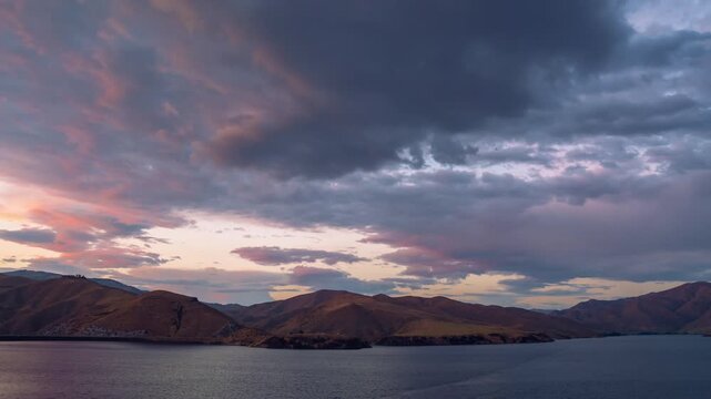 View of the sunset over Lucky Peak Lake with vibrant orange and purple clouds reflecting over the calm water and rolling hills in Ada County, Idaho, United States.