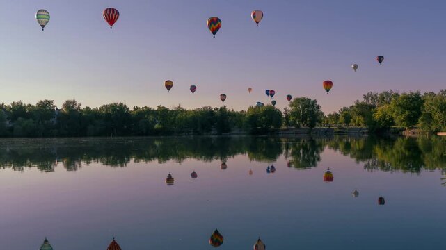 View of colorful hot air balloons floating above a calm lake with clear reflections in the water and trees along the shore in Idaho, United States.