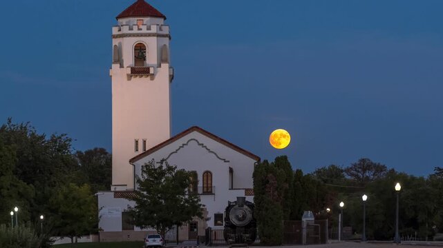 View of historic train depot with a tall bell tower and a steam locomotive parked in front under a large full moon at dusk in Idaho, United States.