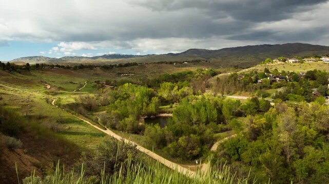 View of Camel's Back Park featuring rolling hills, lush green trees, a small pond, and walking trails under a dramatic cloudy sky in Boise, Idaho, United States.