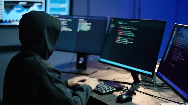 Anonymous hacker in a black hoodie typing computer code on multiple monitors in a dark room. Cybersecurity threat, cybercrime, and digital data hacking concept. Cyber attack and IT programming.