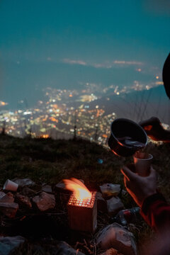Vertical night lifestyle shot of campers pouring a hot drink into a cup over a small portable stove fire. Mountain top viewpoint featuring a blurred city lights background with bokeh.