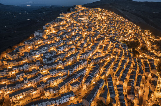 Aerial view of the historic town of Gangi illuminated at night with warm street lights glowing across the dense hillside architecture and narrow streets in Gangi, Sicily, Italy.