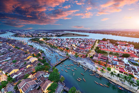 People walk on a bridge as boats float on the Thu Bon River in Hoi An Vietnam during sunset. The sky shows colors as the day ends