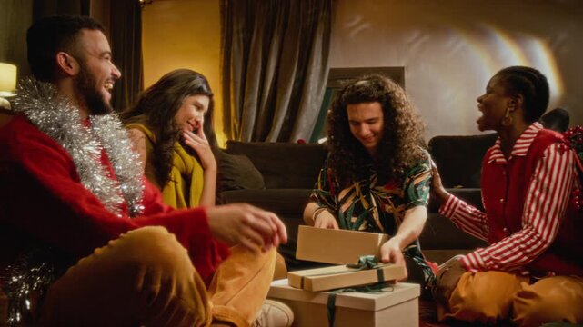 Diverse multiethnic young friends sitting near Christmas tree opening holiday gift box and laughing together. Suitable for festive winter seasonal advertising and retail promotions
