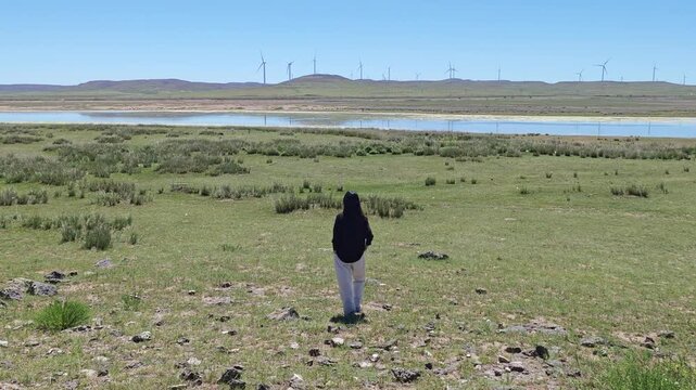 One Asian woman enjoying scenic landscape of salt lake wetland and distant wind turbines in summer in Ulan Hada Volcano Geopark, Ulanqab, Inner Mongolia, China