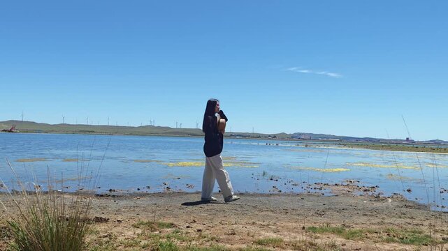 One Asian woman enjoying scenic landscape of salt lake wetland and distant wind turbines in summer in Ulan Hada Volcano Geopark, Ulanqab, Inner Mongolia, China