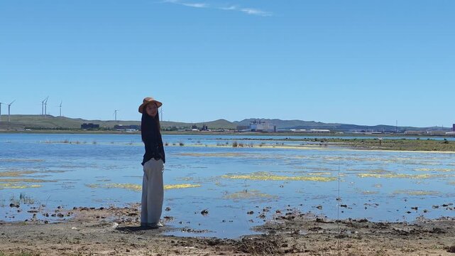 One Asian woman enjoying scenic landscape of salt lake wetland and distant wind turbines in summer in Ulan Hada Volcano Geopark, Ulanqab, Inner Mongolia, China