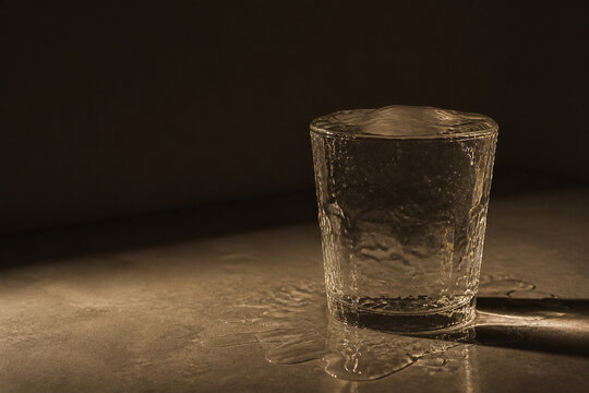 Photorealistic business concept showing a glass on a plain surface overflowing with water running down its sides and pooling around the base, warm side light. Visual metaphor for time pressure and.