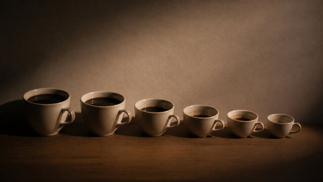 Photorealistic business concept showing a line of used coffee cups of gradually decreasing fill level arranged along a desk edge, warm dim light, plain wall. Visual metaphor for time pressure and.