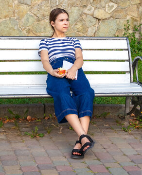Teen girl sitting on a white park bench holding an apple and notebook, looking thoughtful and relaxed
