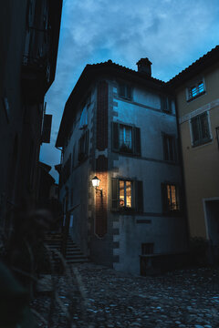 Vertical view of an old stone house in the narrow cobbled streets of Morcote during blue hour. Warm streetlight lantern and illuminated window casting a cozy glow against the deep blue evening sky.