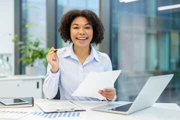 Obraz premium Portrait of a young African American woman sitting at an office desk, holding papers and a pen in her hand and looking at the camera with a smile
