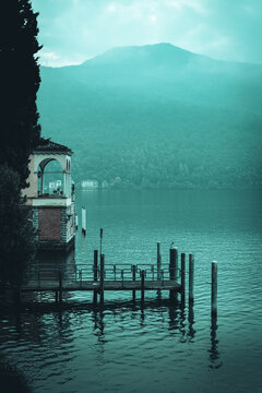 Scenic vertical shot of a traditional stone loggia balcony and wooden pier on the calm waters of Lake Lugano. Misty mountain silhouette and moody teal sky in the background at Morcote, Ticino.