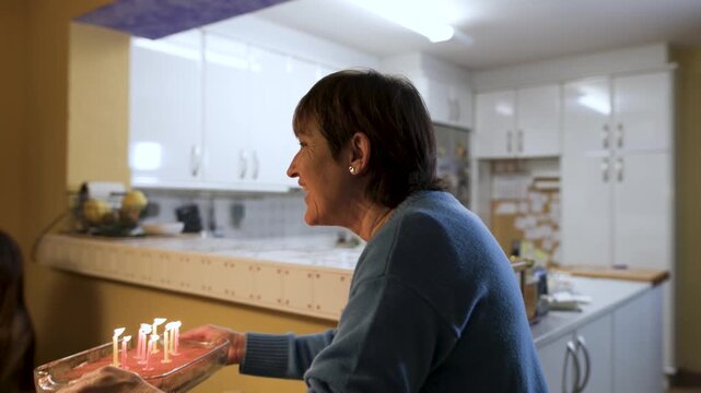 Grandmother carries a tiramisu cake with lit candles to her grandson as family sings and claps. He closes his eyes, makes a wish and blows out candles while everyone applauds. Tracking shot, video 4K