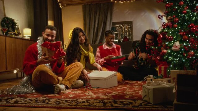 Diverse group of young friends sitting near Christmas tree, unwrapping holiday gift boxes and exchanging presents. Footage ideal for winter retail promotions and seasonal marketing campaigns