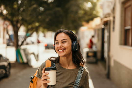 Young woman smiling walking city street with headphones and coffee