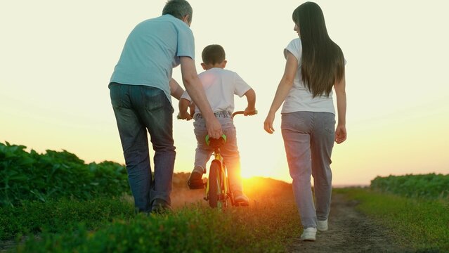Parents teach child to ride bicycle in park. Happy family. Young family with little son riding bicycle. Family with child boy playing at sunset. Teamwork, dad mom son. Sport family. Father mother son