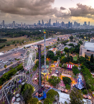 Aerial view of Luna Park amusement park with ferris wheel, drop tower, and roller coaster rides illuminated at dusk, set against the skyline in Tel Aviv-Yafo, Tel Aviv District, Israel.