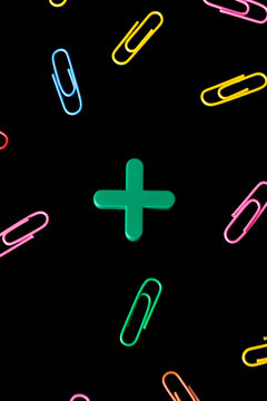 Green wooden plus symbol surrounded by scattered colorful paper clips on black background