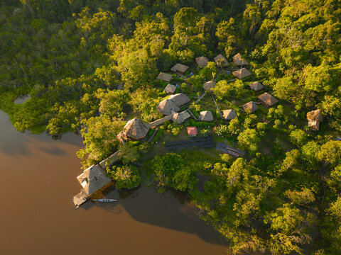 Aerial view of Sani Lodge in the Amazon rainforest, featuring thatched-roof cabins, a wooden walkway, and a river with a boat, in El Retiro, Sucumbios, Ecuador.