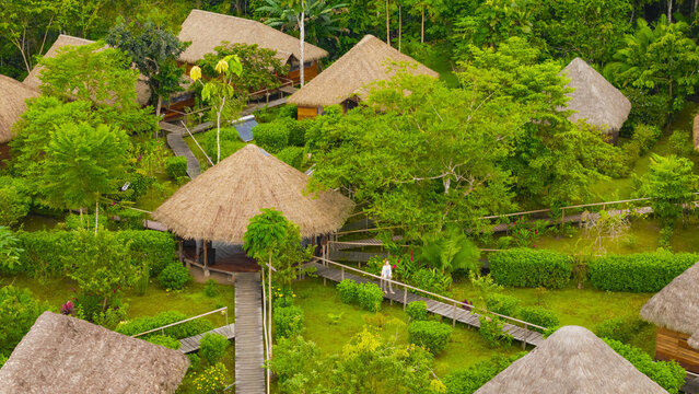 Aerial view of ecolodge featuring thatched-roof cabins in the Amazon rainforest, connected by wooden walkways with a person walking along the path in El Retiro, Sucumbios, Ecuador.