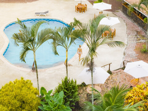 Aerial view of a swimming pool surrounded by palm trees, white umbrellas, and tables with a person walking on the patio in Santa Cruz, Galapagos Province, Ecuador.